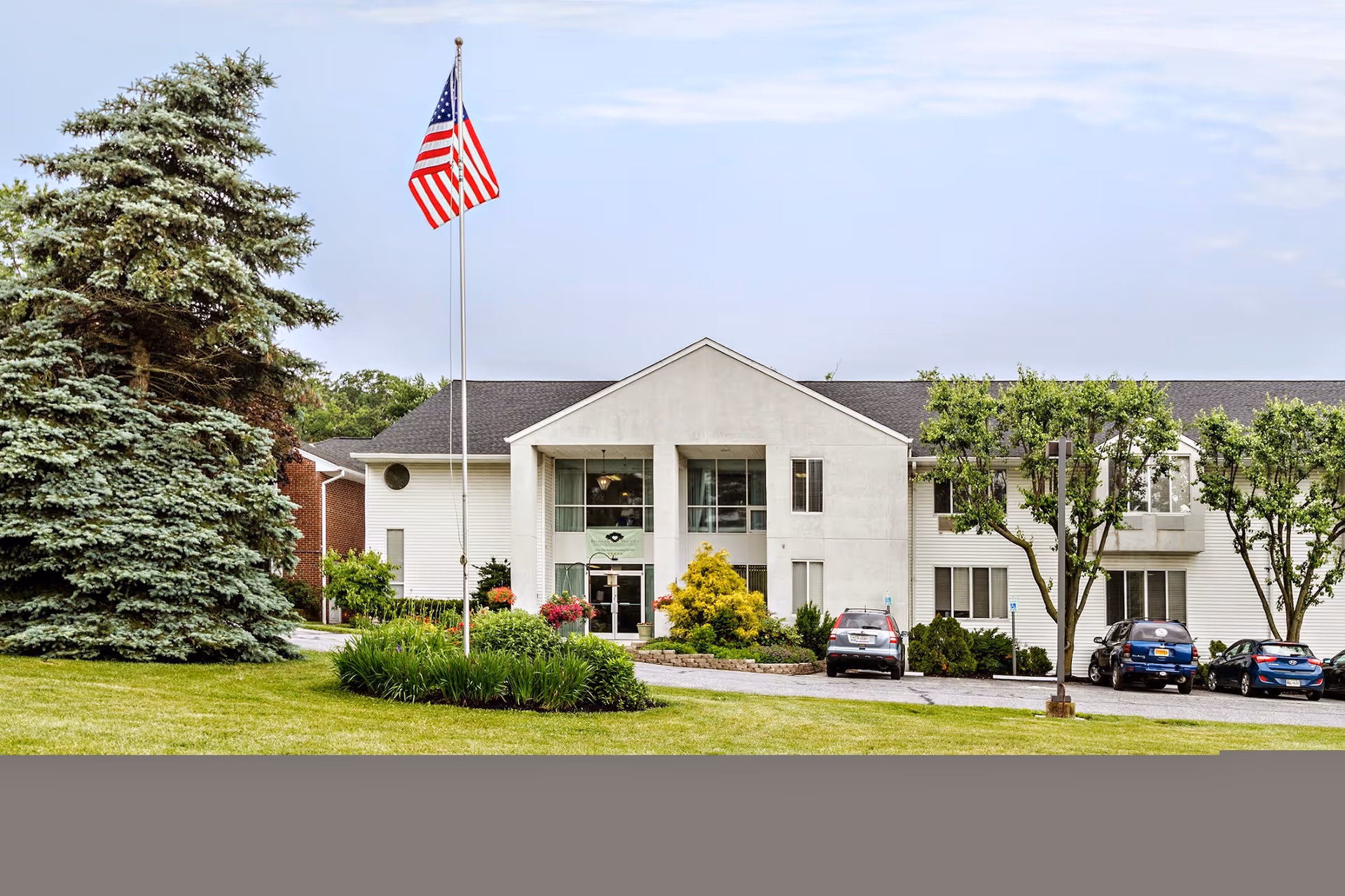 Front exterior of a two-story health and rehabilitation building with an American flag, landscaped lawn, and parked cars.