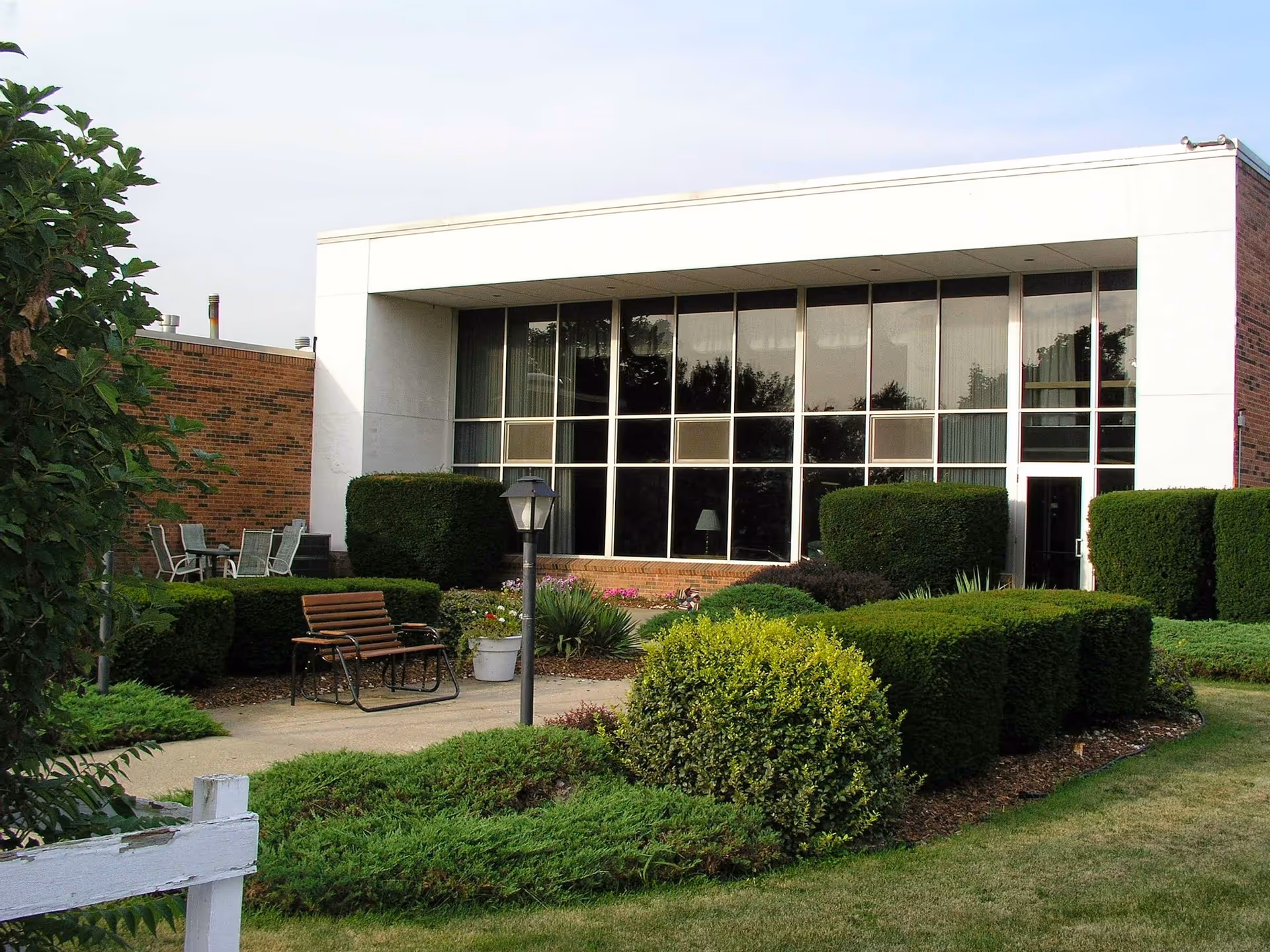 Exterior view of a building with large windows and neatly trimmed bushes and shrubs in front. There is a bench and a lamp post on a paved walkway surrounded by green landscaping.