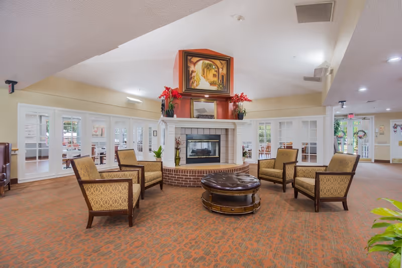 Spacious senior living lounge with armchairs arranged around a central fireplace and round ottoman.