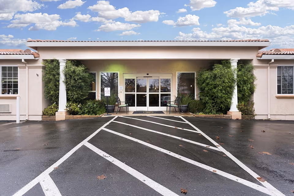 Front entrance of Regency Place Senior Living facility with double glass doors, two white columns, green bushes on either side, and a wet parking lot with white striped markings in front.