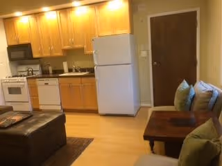 Studio-style interior showing a small kitchen with wood cabinets, refrigerator and stove along the back wall and a seating area with a couch and coffee table in front.
