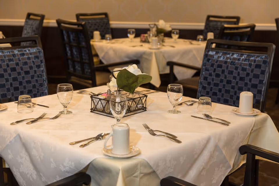 A dining room table set for four with white tablecloth, glassware, silverware, and a centerpiece with a green plant. The chairs have a blue checkered pattern. Additional tables similarly set are visible in the background.
