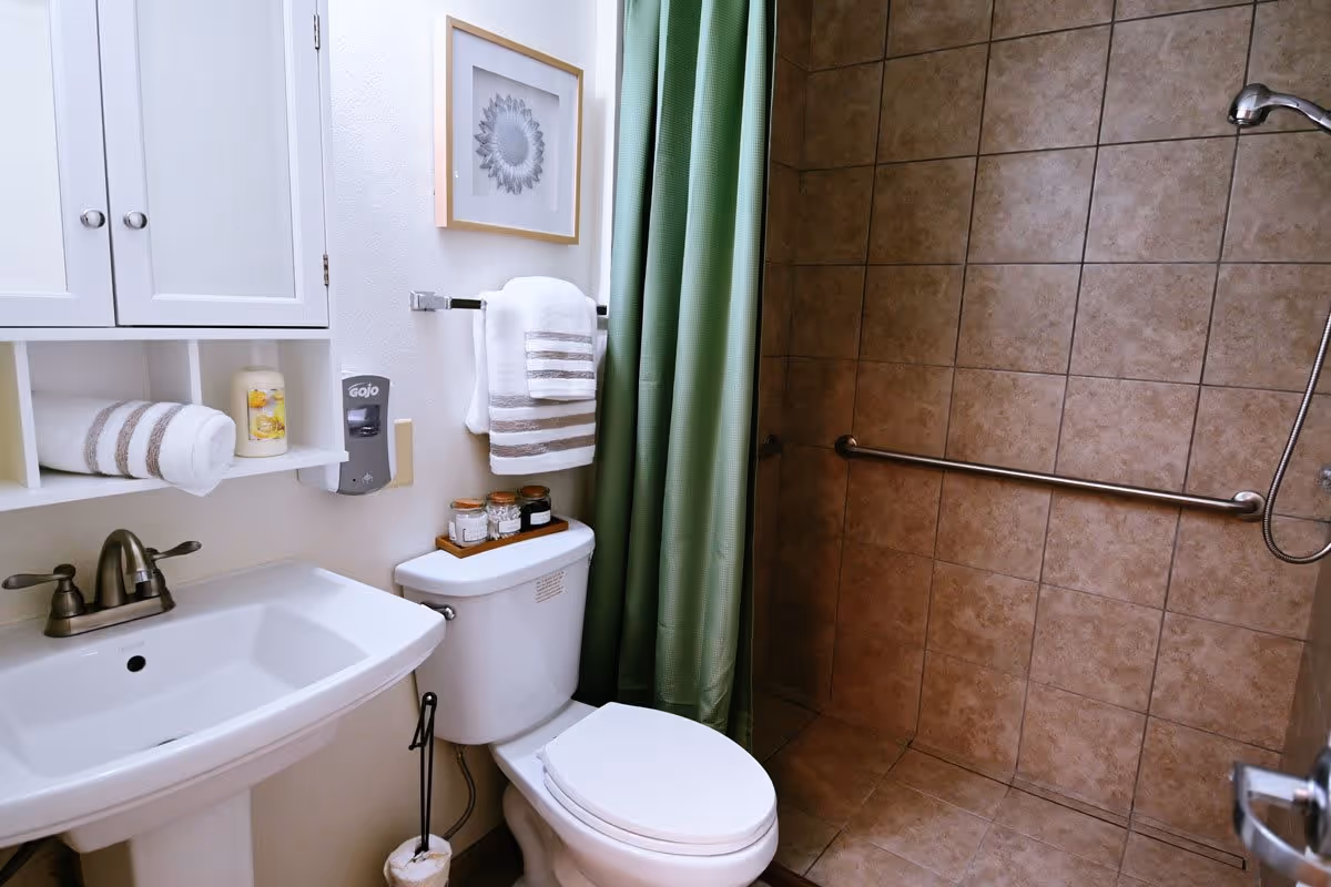 A clean and accessible bathroom with a white pedestal sink, a toilet with a tray holding jars on top, a towel rack with folded white and striped towels, a green shower curtain, and a tiled walk-in shower with a grab bar and handheld showerhead. A white cabinet with shelves and a framed sunflower picture are mounted on the wall.