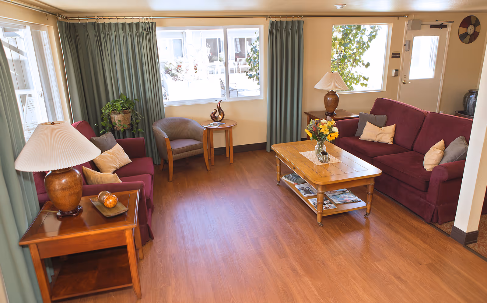 A cozy living room with two maroon sofas adorned with beige and gray pillows, a wooden coffee table with a vase of flowers, two wooden side tables each with a lamp, a small round table with a decorative sculpture, green curtains covering the windows, and a wooden floor.