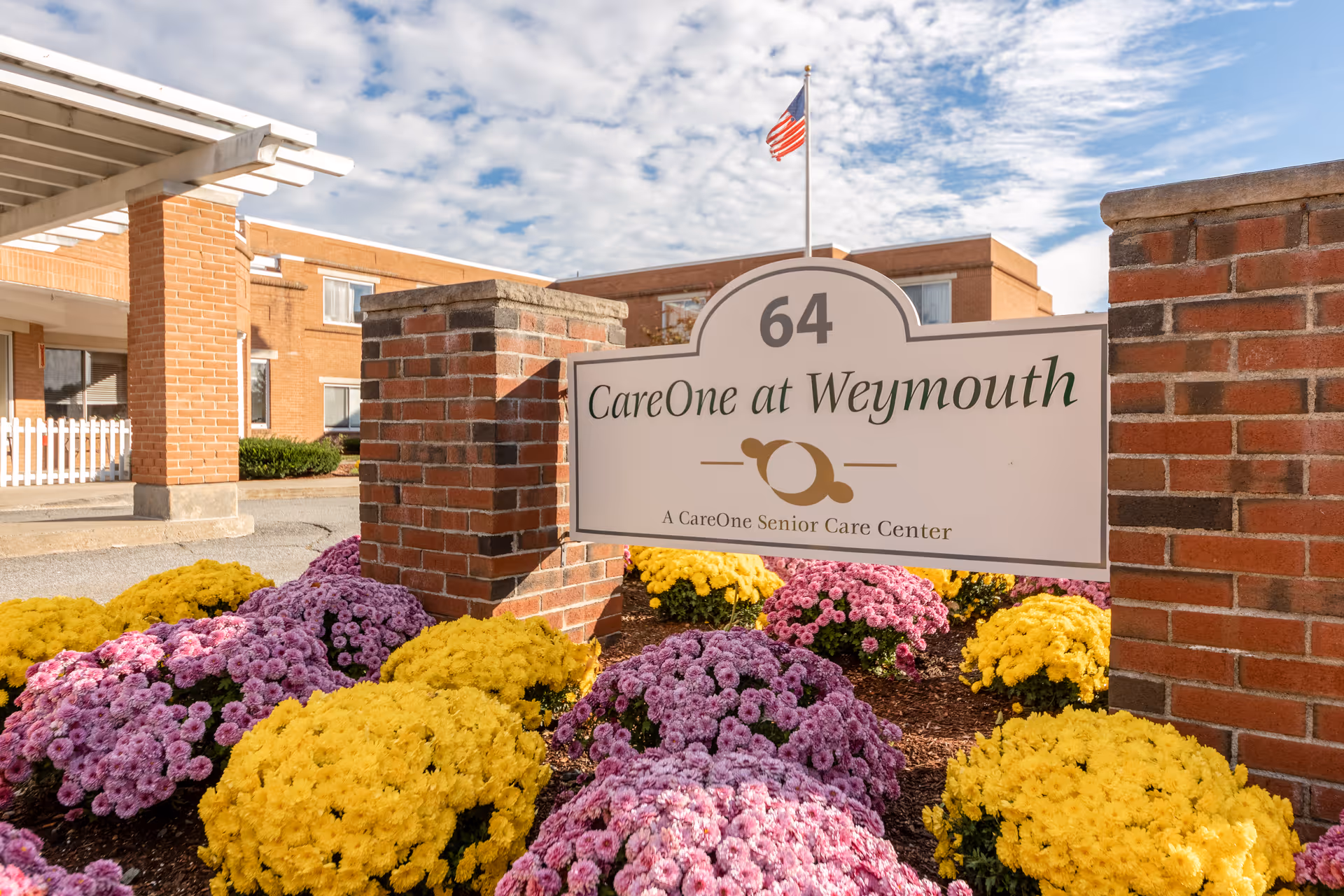 Entrance sign reading 'CareOne at Weymouth' mounted between brick pillars with colorful mums, the facility building, and an American flag in the background.