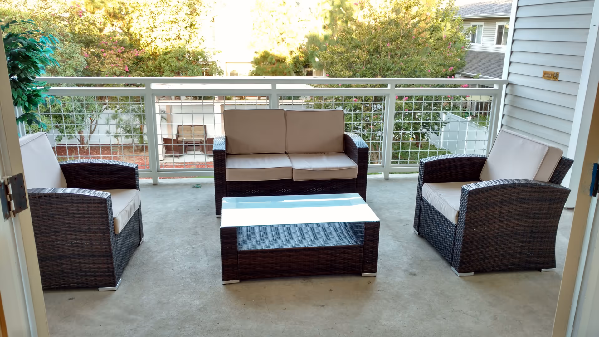 Outdoor balcony area with a set of dark wicker patio furniture including a two-seater sofa, two armchairs with light beige cushions, and a rectangular glass-top coffee table. The balcony has a white railing with a wire mesh design, and there are trees and neighboring buildings visible in the background.