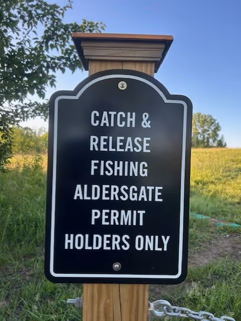 A black and white sign mounted on a wooden post in a grassy outdoor area. The sign reads: 'CATCH & RELEASE FISHING ALDERSGATE PERMIT HOLDERS ONLY'. Trees and a clear sky are visible in the background.