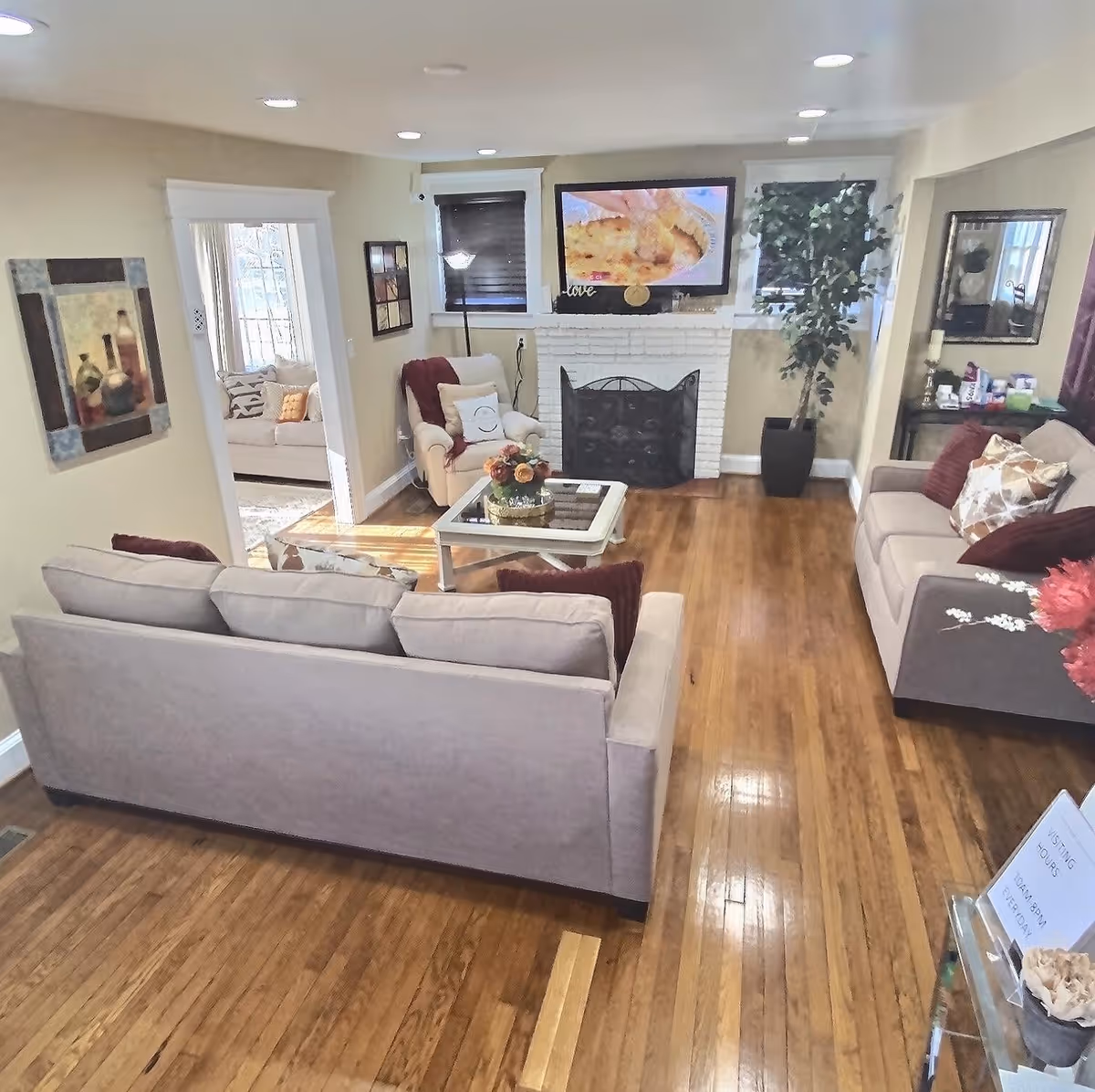 A cozy living room with wooden floors, two beige sofas with pillows, a beige armchair with a red throw, a glass coffee table with a flower arrangement, a white brick fireplace with a TV mounted above it, two windows with dark blinds, a tall floor lamp, a large potted plant, and wall decorations including a mirror and framed pictures.