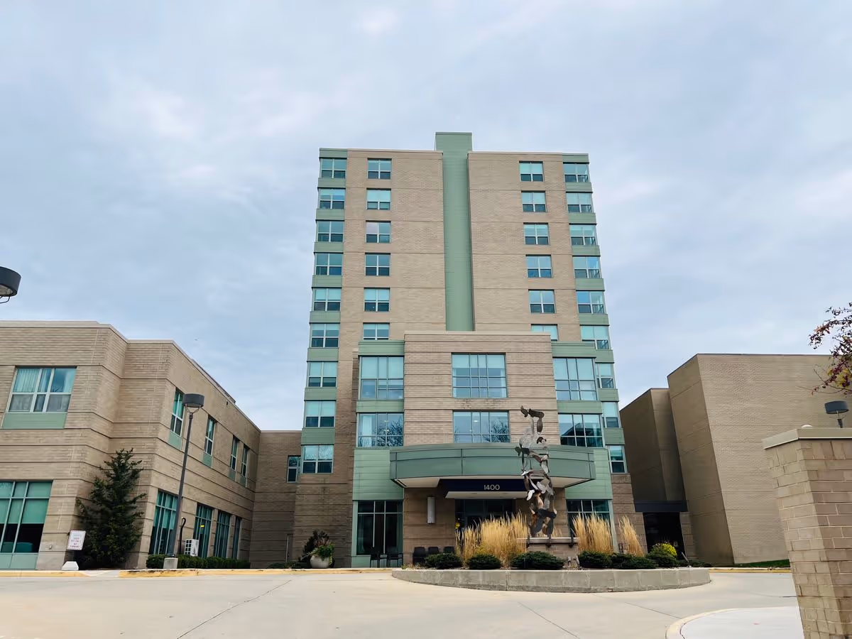 Front view of a multi-story senior living building with a sculpture and driveway at the entrance under a cloudy sky.