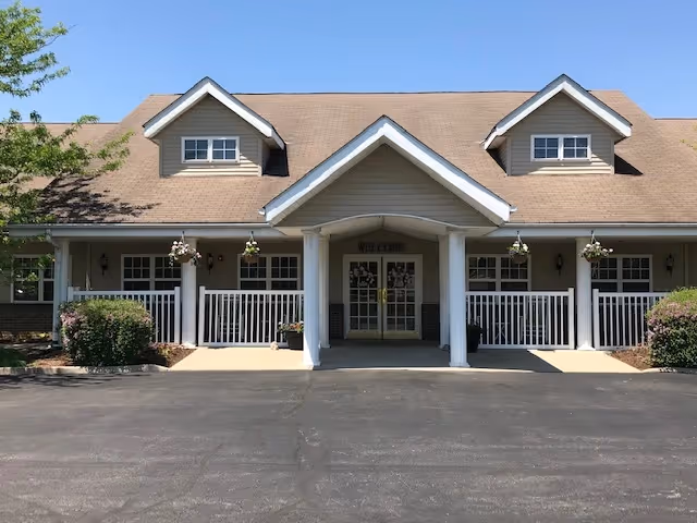 Front exterior view of a single-story building with a peaked roof and two dormer windows. The entrance has a covered porch supported by white columns, with a 'WELCOME' sign above the double glass doors. There are hanging flower baskets and bushes on either side of the porch, and a paved driveway in front.