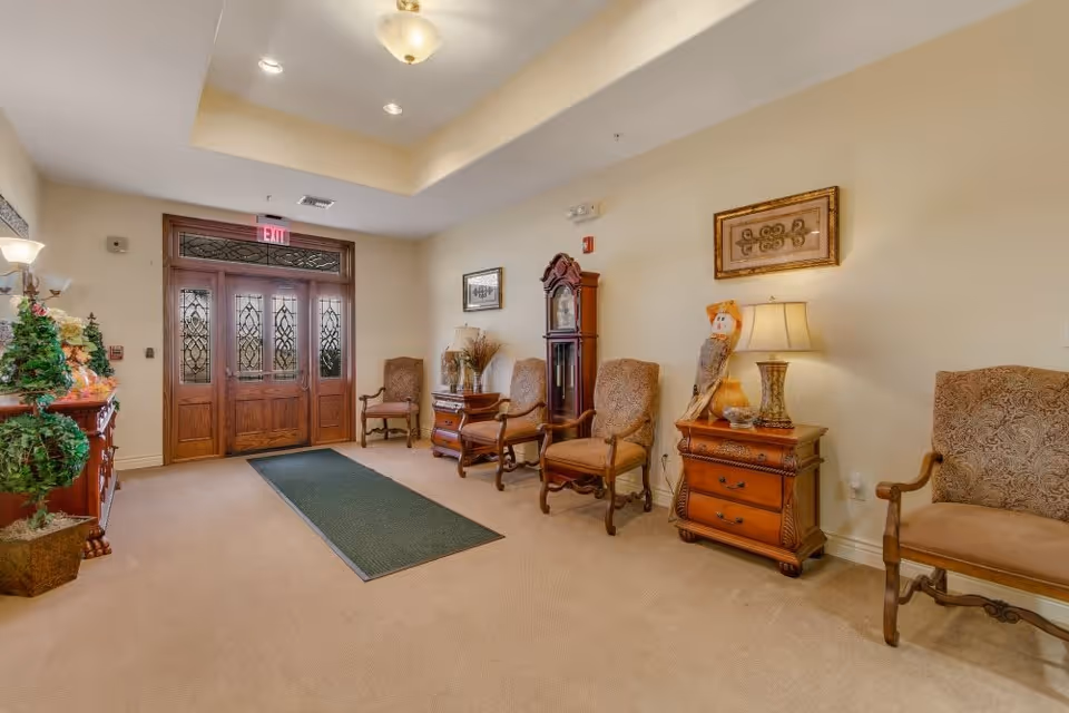 A well-lit interior hallway with beige walls and carpet, featuring a row of wooden chairs with upholstered seats and backs along the right wall. There is a wooden grandfather clock and a side table with a lamp and decorative items. On the left side, there is a wooden cabinet with plants and decorations. At the end of the hallway are double wooden doors with decorative glass panels and an exit sign above them.