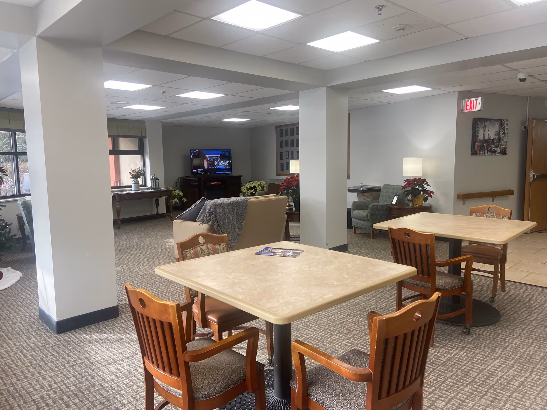 A common area in a senior living facility with two square tables each surrounded by four wooden chairs with cushions. In the background, there is a seating area with a beige couch, a green armchair, a TV on a stand, and several potted plants. The room has carpeted flooring, white walls, and a drop ceiling with fluorescent lights. An exit sign is visible above a door on the right side.
