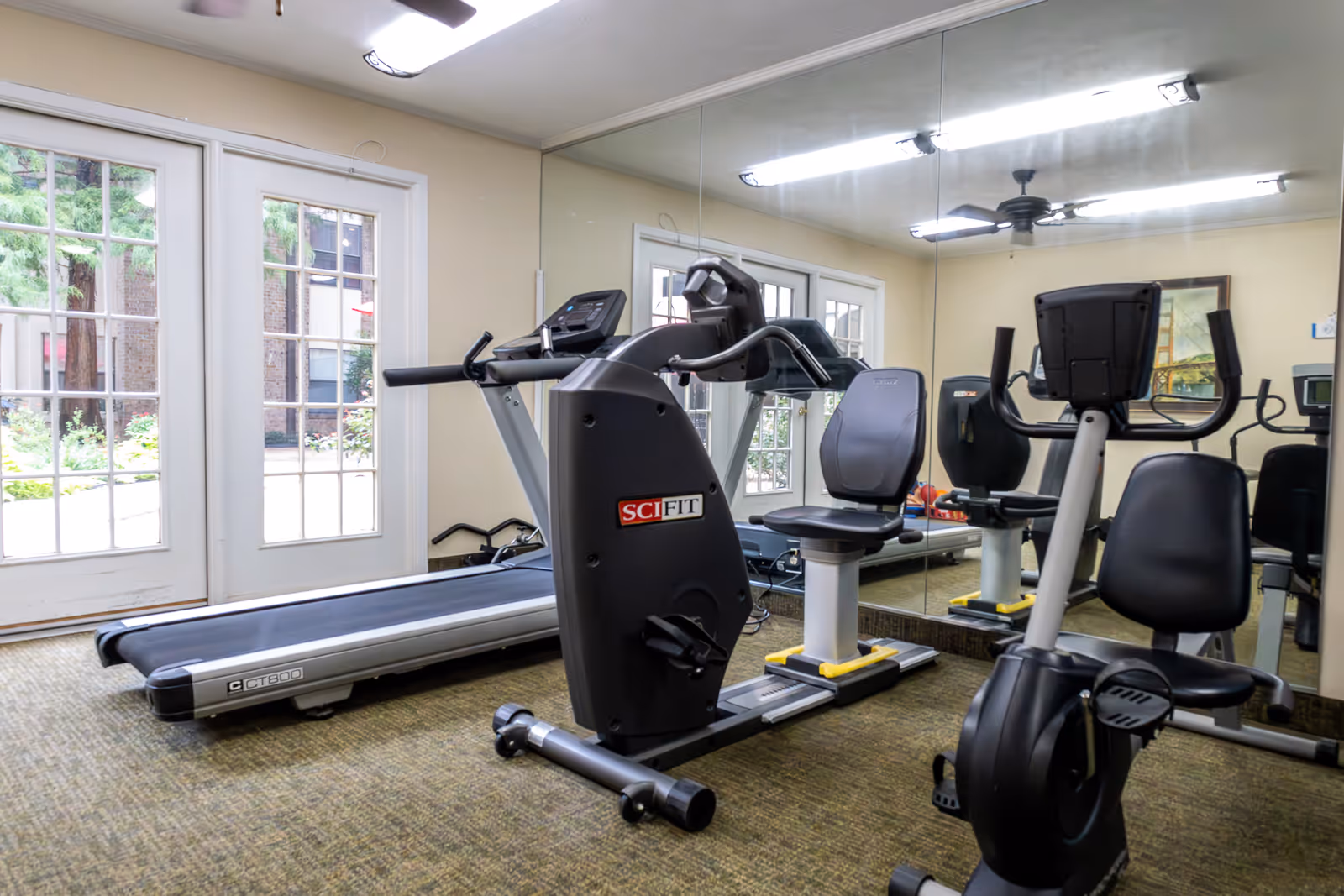 A small fitness room with exercise equipment including a treadmill, a recumbent bike, and an upright stationary bike. The room has large windows and glass doors letting in natural light, and a large mirror covering one wall reflecting the equipment and part of the room.