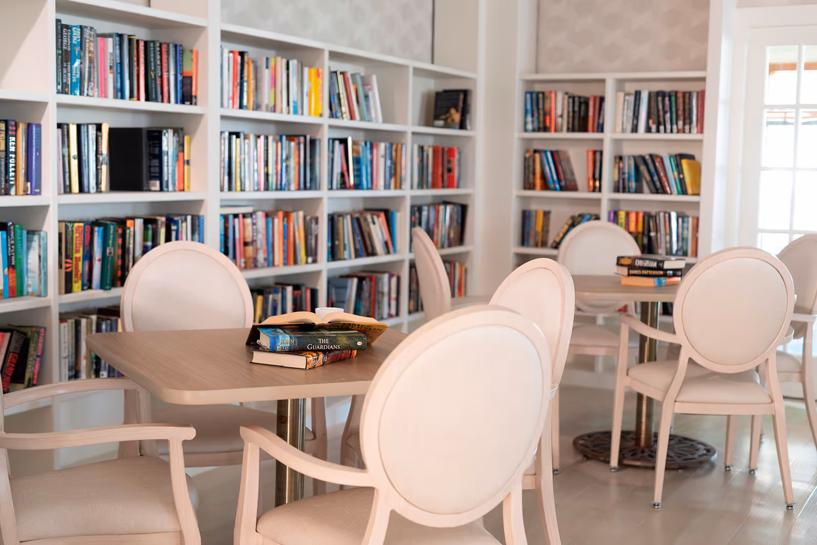 A cozy library room with white bookshelves filled with books lining the walls. There are light-colored wooden tables and cushioned chairs arranged for reading or small group gatherings. Some books are stacked on the tables.