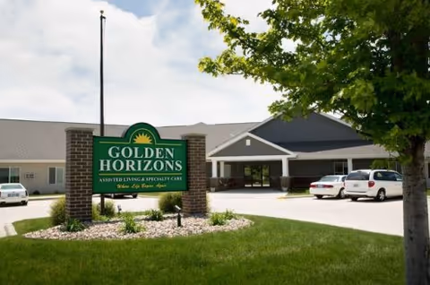 Exterior view of Golden Horizons assisted living facility showing the main entrance, a green sign with the facility name, a flagpole, parked cars, and a tree on a sunny day.