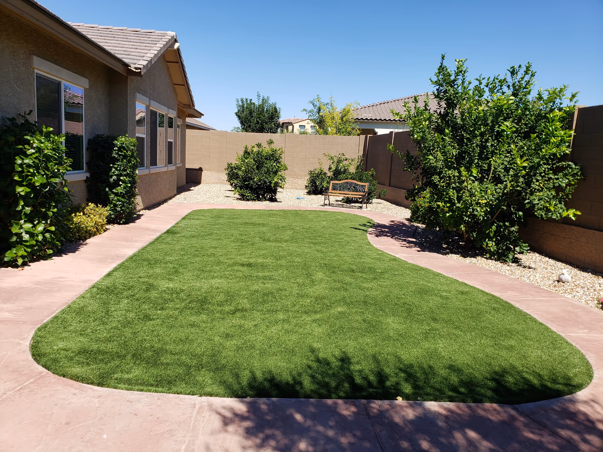 A sunny outdoor garden area with a well-maintained green lawn surrounded by a paved walkway. There are bushes and small trees along the edges, a beige stucco building with windows on the left, and a wooden bench near the back wall under clear blue sky.