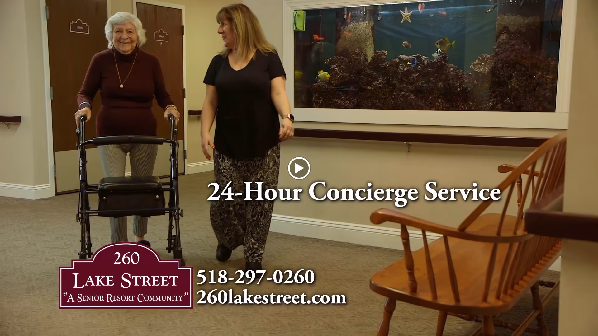 An elderly woman using a walker is accompanied by a caregiver walking down a hallway with doors and a large aquarium mounted on the wall. There is a wooden bench on the right side of the hallway.