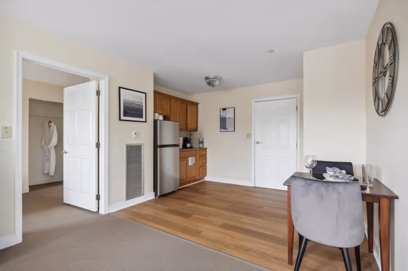 Interior view of a senior living facility apartment showing a small kitchenette with wooden cabinets and a stainless steel refrigerator. To the right, there is a small wooden desk with a gray chair, a wine glass, and a decorative bowl. On the left, an open door reveals a closet with a white robe hanging inside. The floor transitions from carpet to wood, and the walls are painted light beige with framed artwork and a large wall clock.