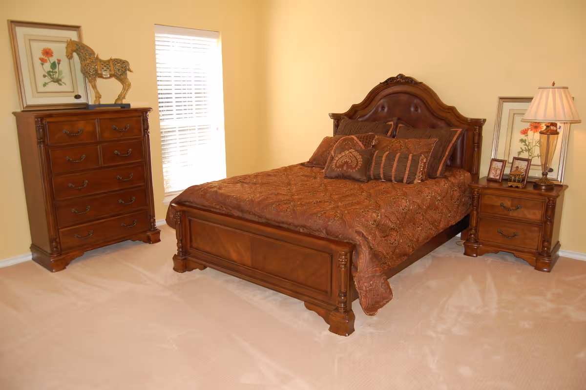 A bedroom with a wooden bed frame and headboard, covered with a patterned brown bedspread and multiple pillows. To the right of the bed is a wooden nightstand with a lamp, framed pictures, and a floral artwork behind it. To the left of the bed is a wooden chest of drawers with a decorative horse sculpture and a framed floral picture on top. The room has beige carpet and light yellow walls with a window letting in natural light.