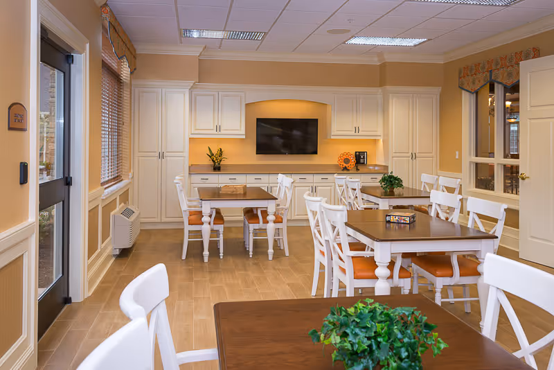 A bright and clean dining room with several wooden tables and white chairs with orange cushions. The room features a wall-mounted flat screen TV, white cabinetry, and decorative plants on the tables. Large windows with blinds and a glass door provide natural light.