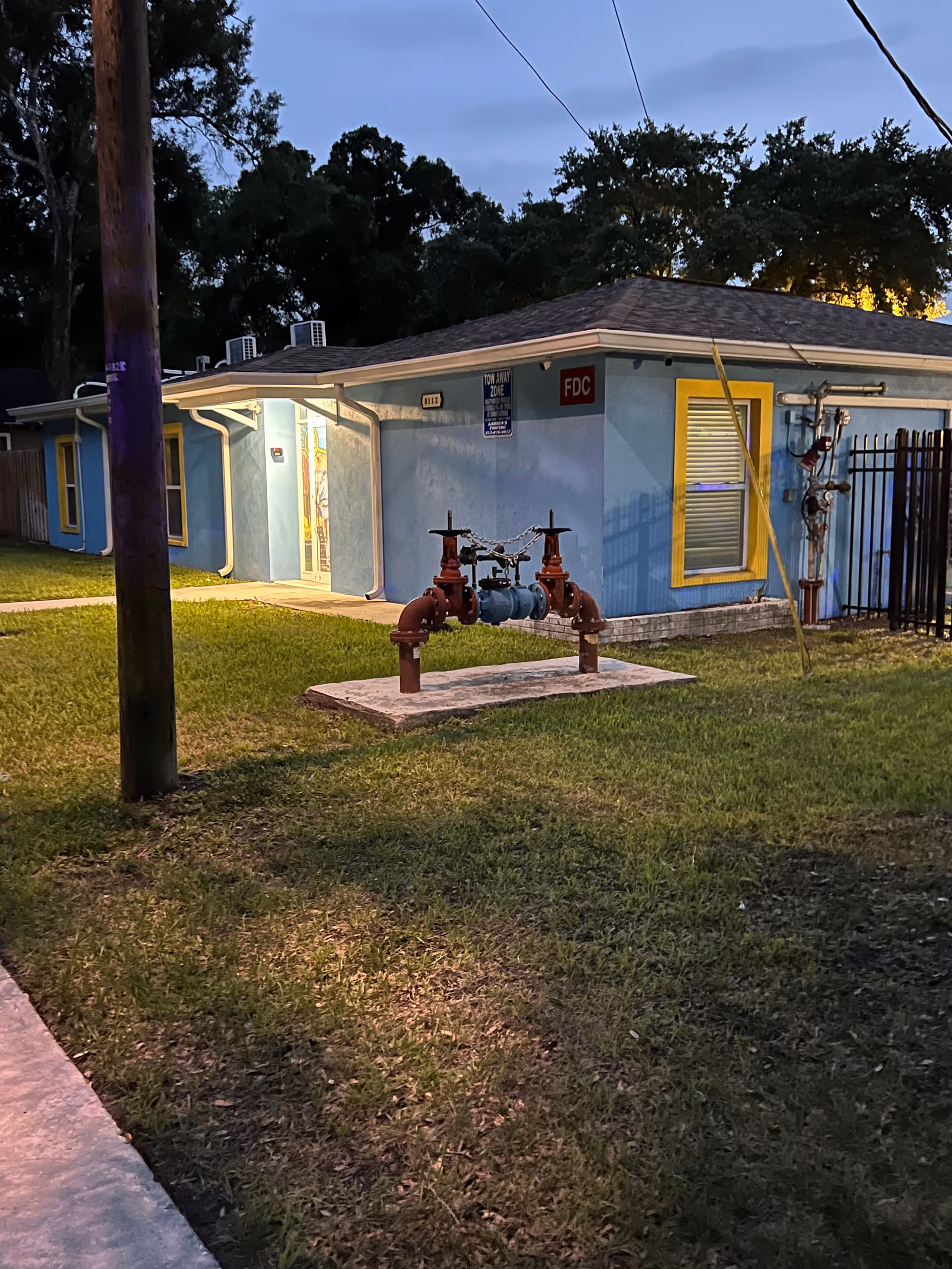 Exterior view of a single-story blue building with yellow window frames at dusk. There is a grassy area in front with a red fire department connection pipe assembly on a concrete slab. A wooden utility pole is visible on the left side, and the building has a covered entrance with a light on.