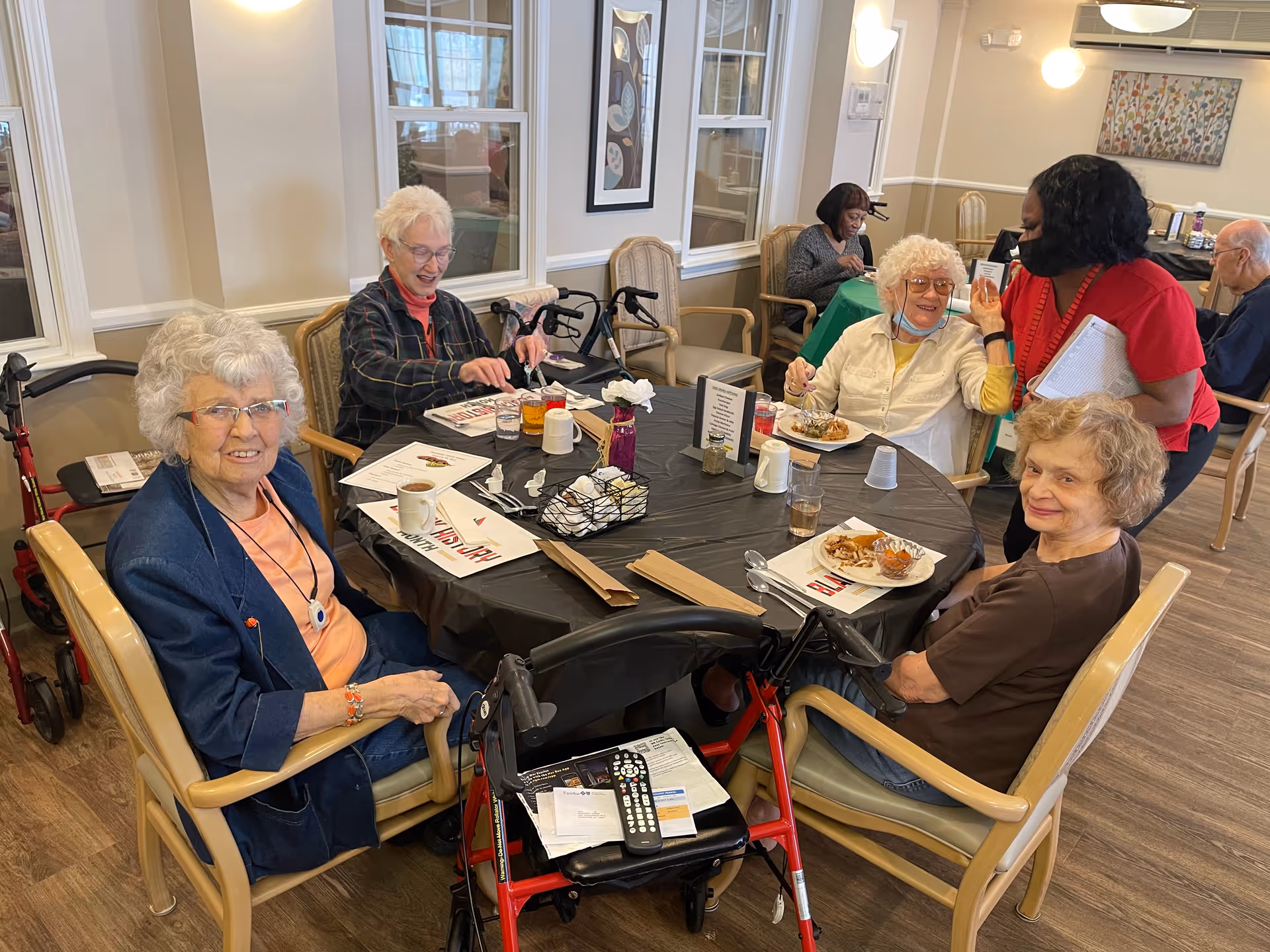 A group of elderly residents sitting around a table in a dining room, eating and chatting with a staff member.