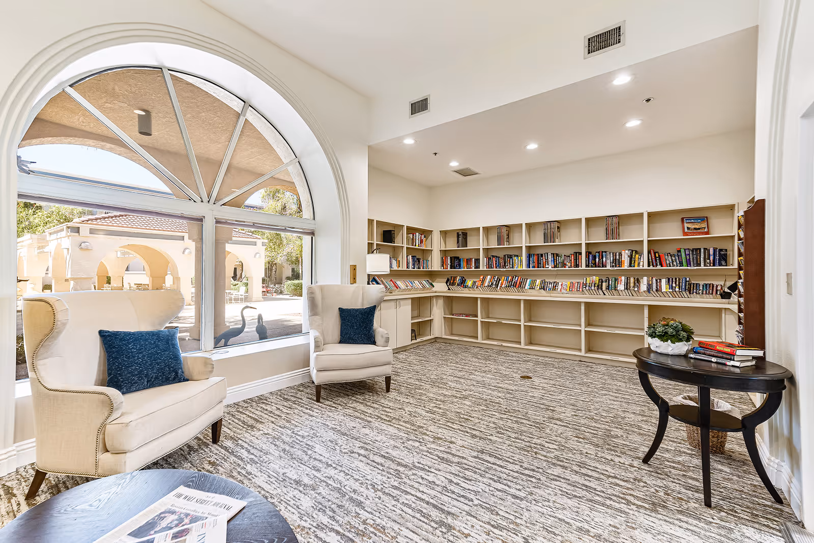 A bright and cozy reading room in a senior living facility with large arched windows letting in natural light. The room features two beige armchairs with blue cushions, a round wooden table with newspapers, built-in bookshelves filled with books, and a small side table with a plant and more books. Outside the window, there is a view of an outdoor patio area with arches and greenery.