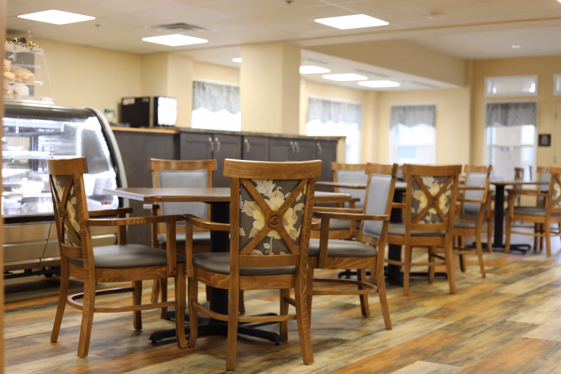 A dining area with wooden tables and chairs featuring floral patterned backs. The room has large windows with curtains, a display case with baked goods, and cabinets along the back wall. The flooring is wood-style and the lighting is bright with ceiling fixtures.
