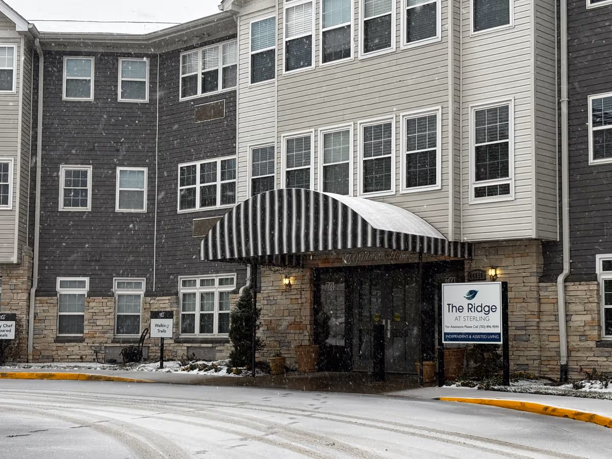 Front entrance of a multi-story senior living building with a striped canopy, an entrance sign reading "The Ridge at Sterling", and light snowfall.