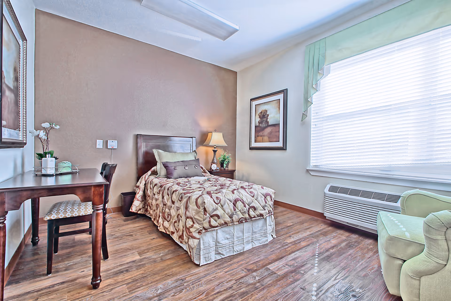 A cozy bedroom with a single bed covered in a patterned quilt, a wooden desk with a chair, a bedside table with a lamp and plant, a framed picture on the wall, a large window with blinds and a green valance, and a green upholstered armchair on wood flooring.