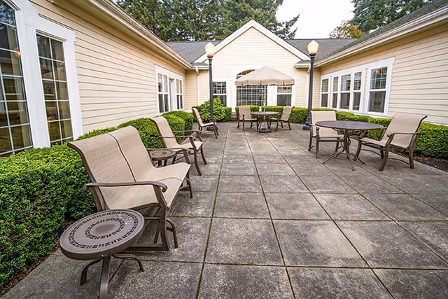 Outdoor patio area at Brookdale Olympia West with several cushioned chairs and tables arranged on a tiled floor, surrounded by beige building walls and green shrubbery, under a partly cloudy sky.