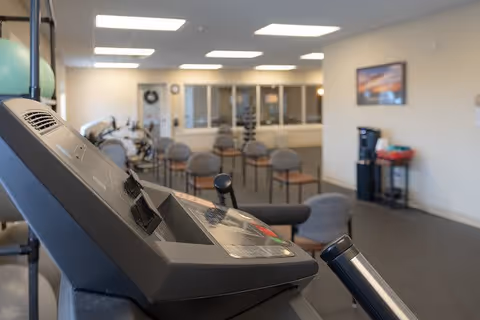 Interior view of a fitness room in a senior living facility with exercise equipment in the foreground and several chairs arranged in rows in the background. The room has bright overhead lighting, a water dispenser, and a framed picture on the wall.