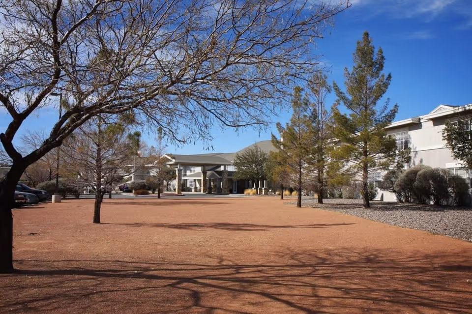 View of the exterior grounds of a senior living facility with a large open dirt area, leafless trees, and a building in the background under a clear blue sky.
