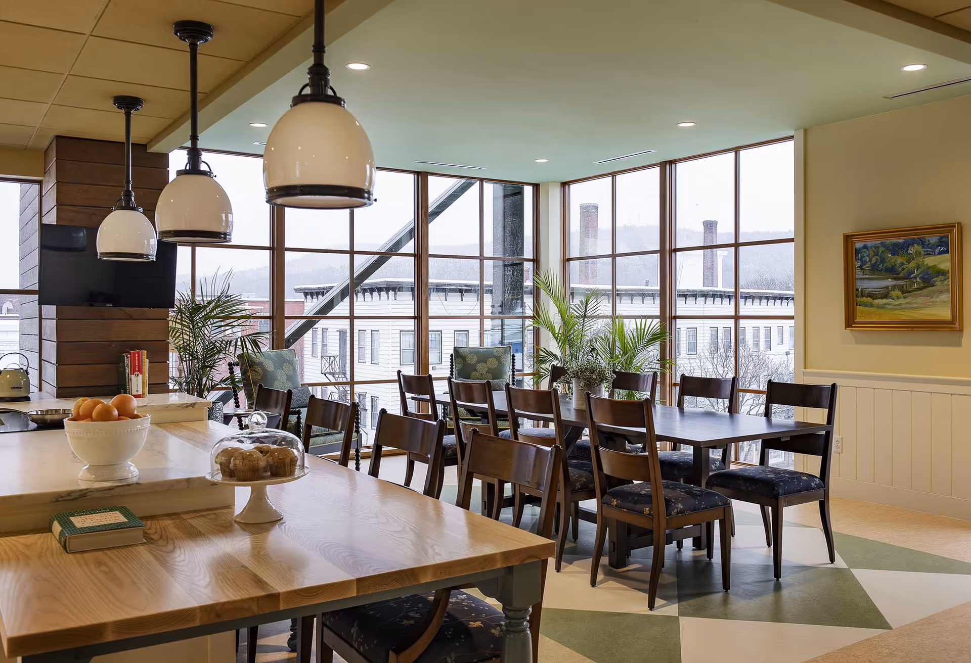 A bright dining area with large windows showing an exterior view of buildings and hills. The room features a long wooden dining table with multiple chairs, a kitchen island with a bowl of oranges and a cake stand with muffins, three hanging pendant lights, potted plants, a wall-mounted TV, and a framed landscape painting on the wall.