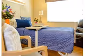 A bright and cozy bedroom in a senior living facility featuring a bed with a blue quilt and blue pillows, a wooden chair with a white cushion, a small table with a lamp and flowers, and a large window letting in natural light.