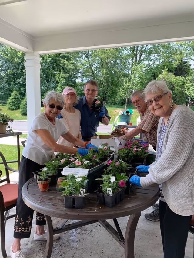 Five elderly individuals standing around a round outdoor table filled with potted flowering plants, engaging in a gardening activity under a covered patio with green trees and grass visible in the background.