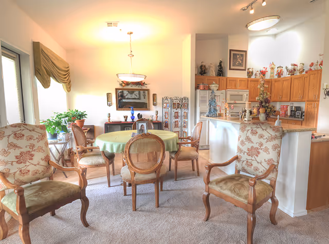 A bright and cozy senior living community common area featuring a round dining table with a green tablecloth surrounded by six wooden chairs with cushioned seats. The room includes two upholstered armchairs with floral patterns in the foreground. The background shows a kitchen area with wooden cabinets, a refrigerator, microwave, and various decorative items on the countertops and above the cabinets. There are plants near the windows and a decorative mirror on the wall above a small cabinet.