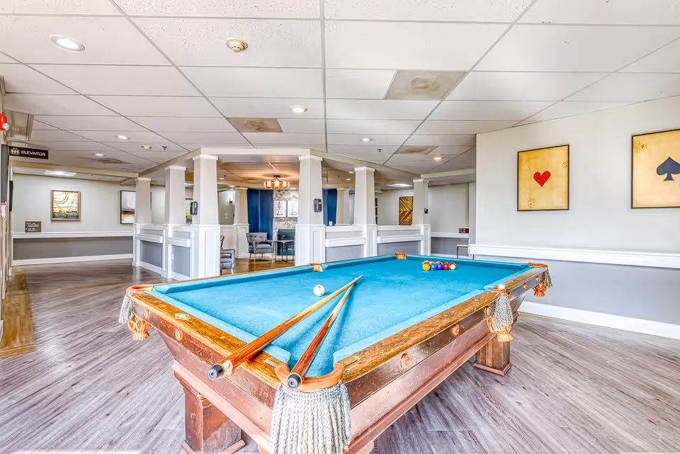 Interior view of a recreational room at Sellwood Senior Living featuring a pool table with two cues resting on it. The room has light-colored walls with framed artwork of playing card suits, wood flooring, and a seating area with chairs and a table in the background.