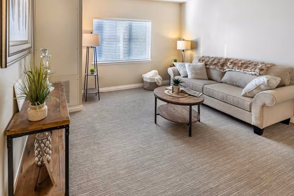 Neutral-toned living room featuring a beige sofa with throw pillows and blanket, round wooden coffee table, console table with plants, and two floor lamps by a window.
