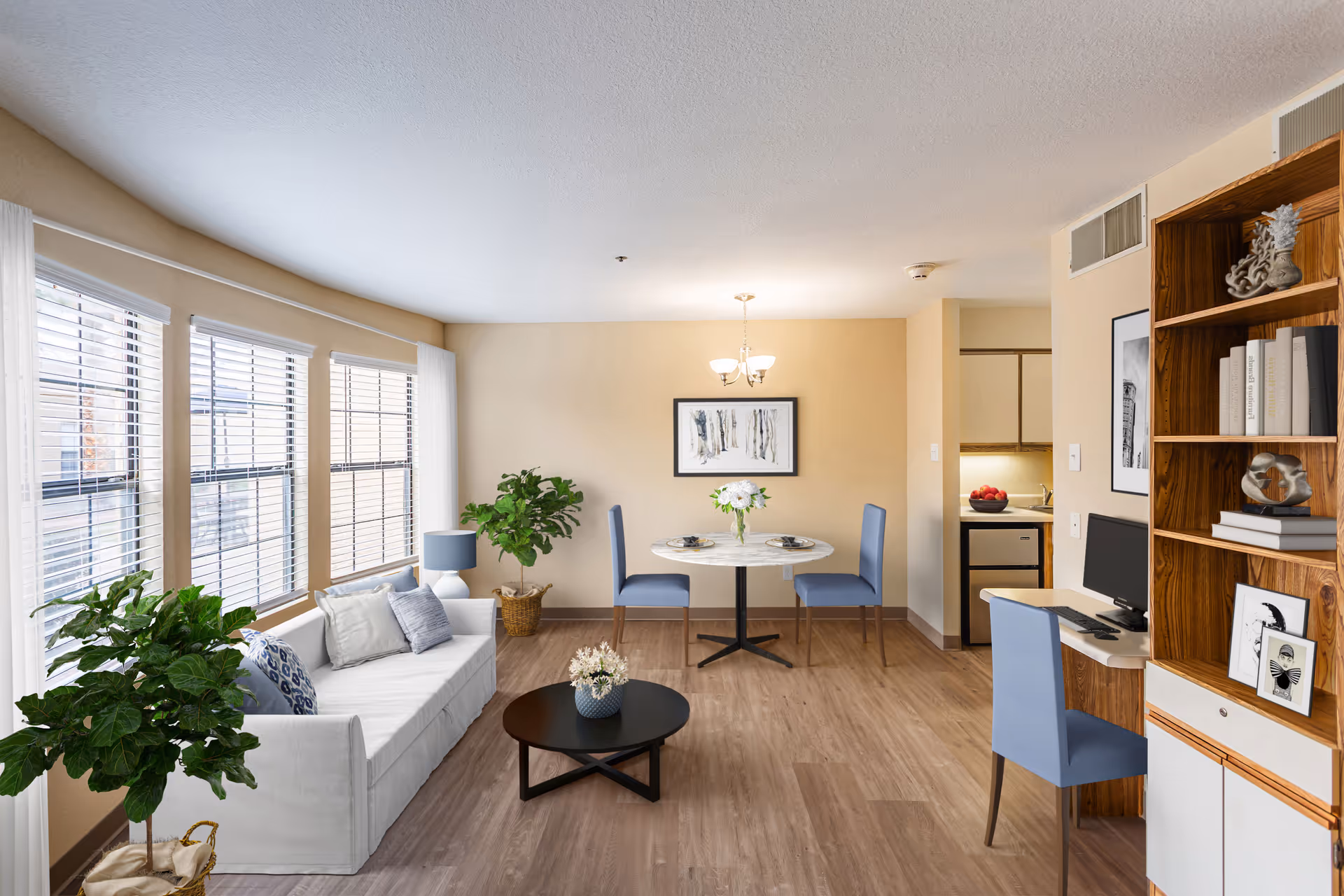 A bright and cozy living area in a senior living facility featuring a white sofa with cushions, two green potted plants, a round black coffee table with a flower vase, a small round dining table with two blue chairs and a flower centerpiece, a wooden bookshelf with books and decorative items, and a small desk with a computer and a blue chair. Large windows with white blinds allow natural light to fill the room.