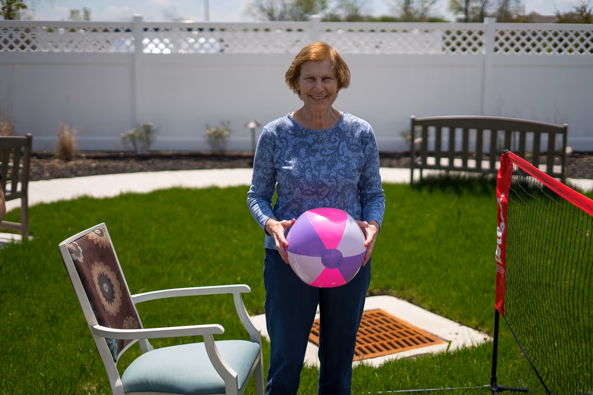 An elderly woman standing outdoors on green grass holding a pink and purple beach ball. She is smiling and wearing a blue patterned long-sleeve shirt and dark pants. There is a white fence in the background, along with a bench, a chair, and a red net set up on the grass.