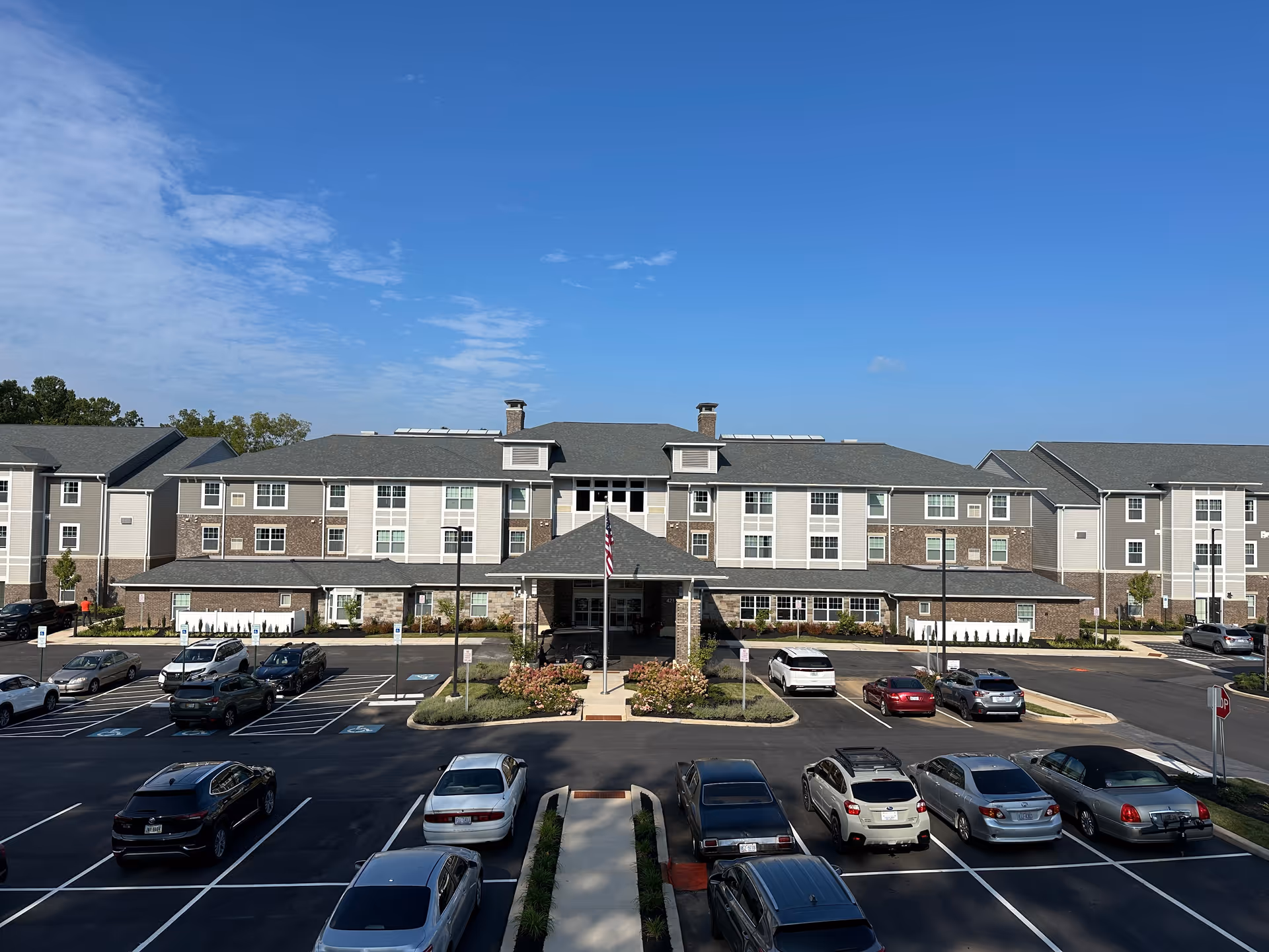 Front exterior view of Julian Woods Retirement Community, a large three-story building with a covered entrance, multiple windows, and a parking lot with several parked cars in front. The sky is clear and blue.