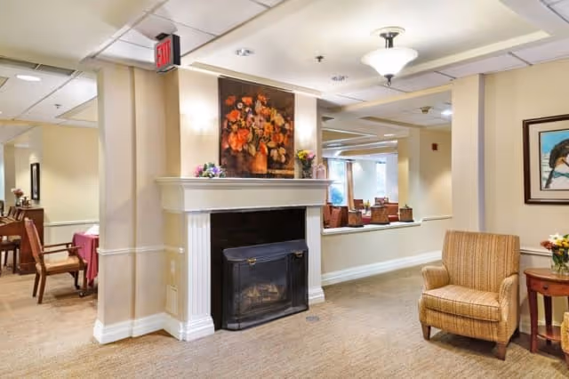 A cozy interior common area featuring a fireplace with a floral painting above it, a comfortable armchair, and a small side table with a flower arrangement. The space has beige walls, carpeted floors, and ceiling lights, with a dining area visible in the background.