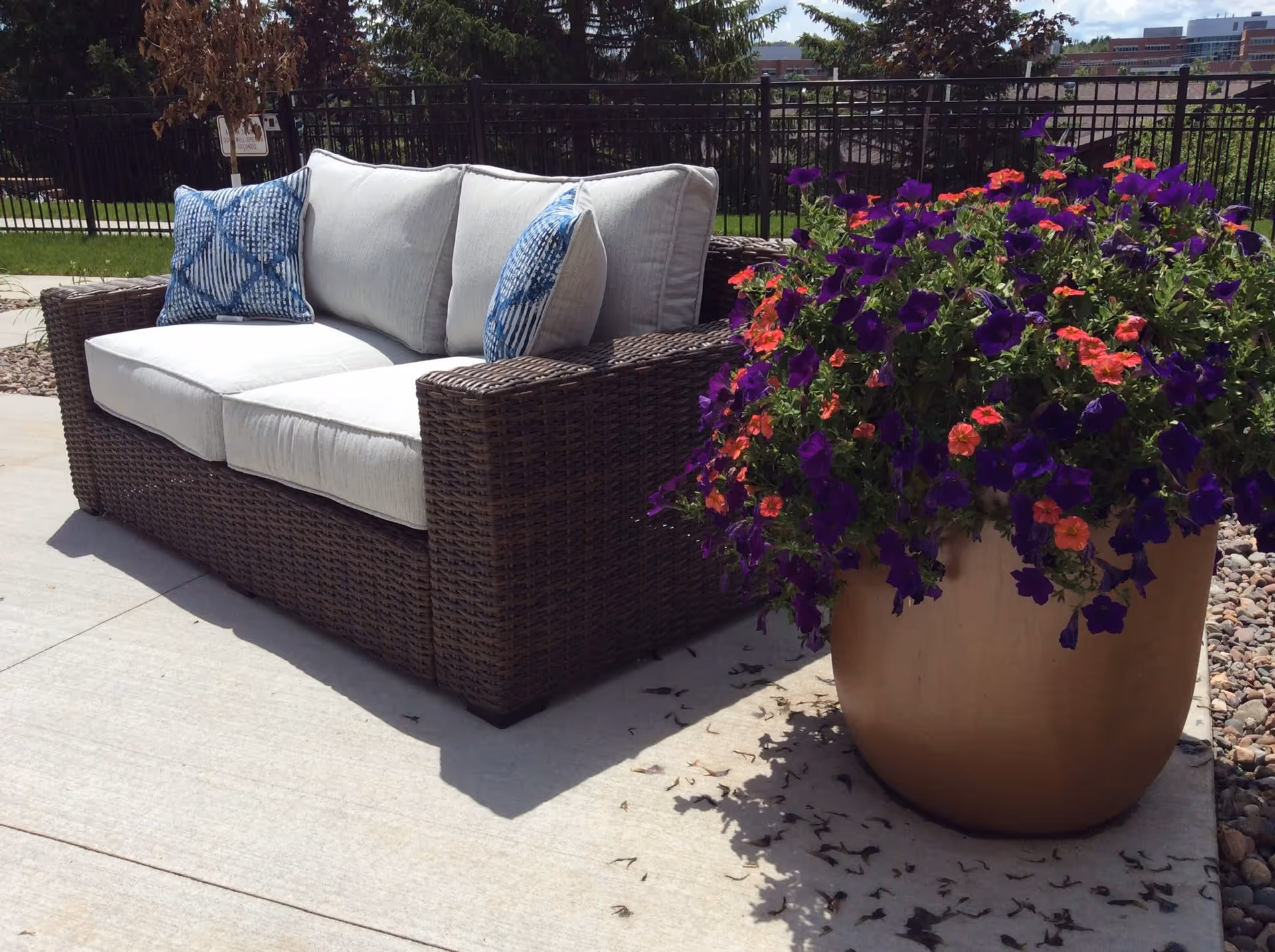 Outdoor patio area with a brown wicker sofa featuring white cushions and two blue patterned pillows, next to a large pot filled with vibrant purple and orange flowers. A black metal fence and green grass are visible in the background under a partly cloudy sky.