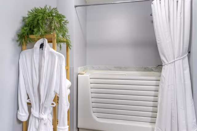 A walk-in bathtub with a white shower curtain tied to the side, next to a wooden shelf holding a white bathrobe and a green potted plant on top.