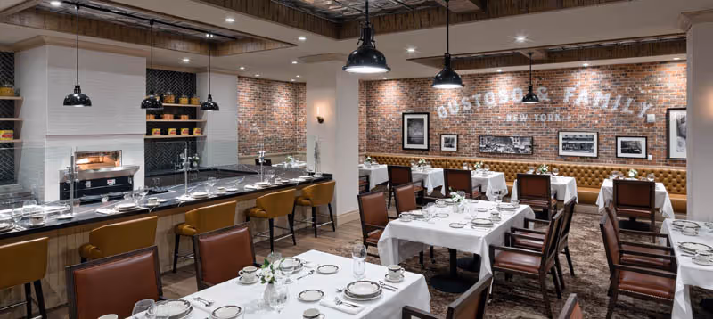 A stylish dining room with white‑clothed tables, leather chairs, a serving counter and exposed brick walls.