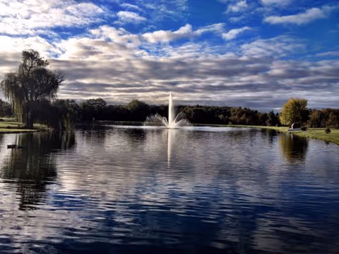 A serene outdoor scene featuring a large pond with a central water fountain spraying upwards. The pond is surrounded by grassy banks with trees, including a large willow tree on the left. The sky above is partly cloudy with patches of blue visible.