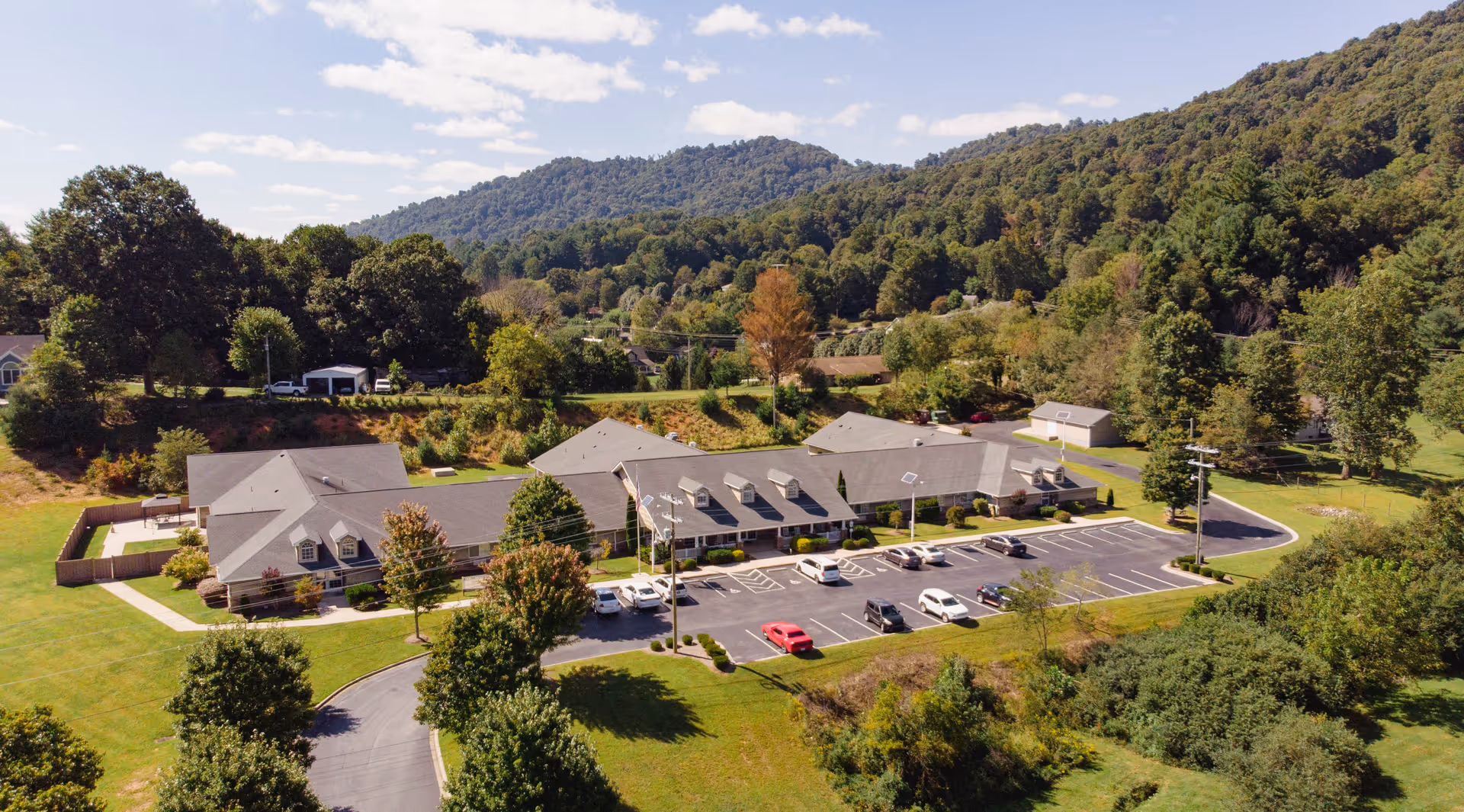 Aerial view of Yancey House, a single-story senior living facility surrounded by greenery and trees, with a parking lot in front and mountains in the background under a partly cloudy sky.