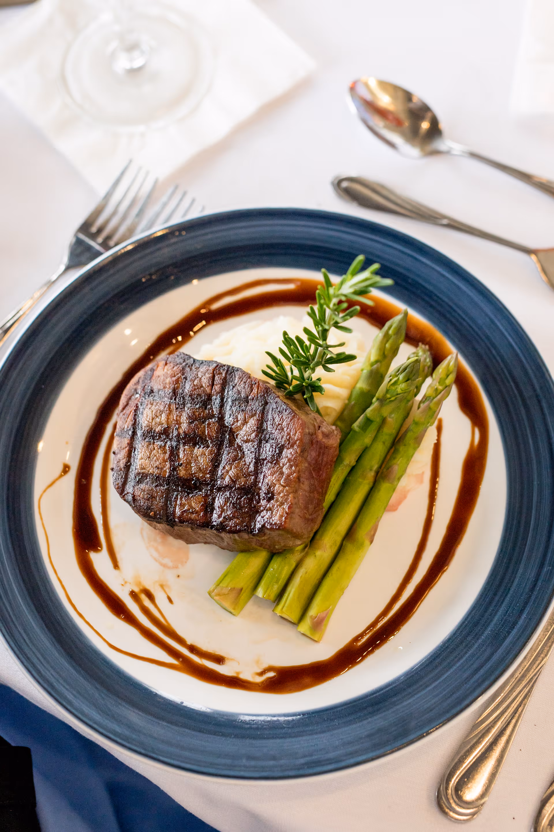 Plated grilled steak with asparagus, mashed potatoes and a balsamic-style sauce on a blue-rimmed plate at a dining table.