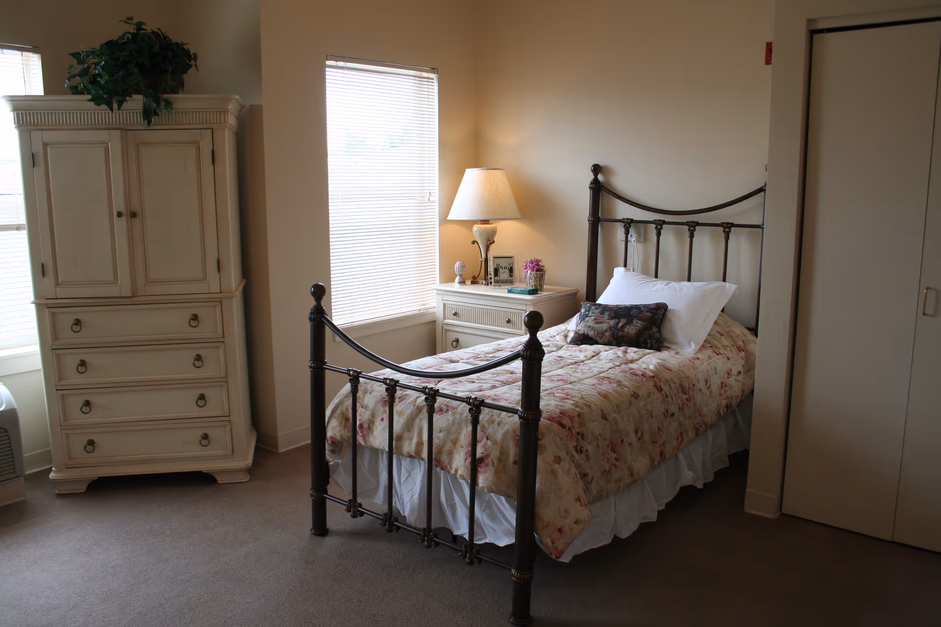 A cozy bedroom with a single metal bed frame, floral bedspread, and a decorative pillow. Next to the bed is a white nightstand with a lamp, a small plant, and framed photos. There is a white dresser with drawers and a cabinet, topped with a green leafy plant. The room has beige walls, carpeted floor, and two windows with blinds allowing natural light in.